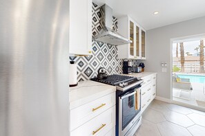 A modern kitchen with white cabinets, a stainless steel range hood, and a large window overlooking a pool area. - Mid-century modern hacienda with pool, outdoor kitchen, & fireplace - dogs ok (Palm Springs)