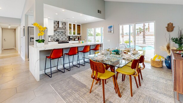 Una cocina moderna con una isla central, taburetes de bar y un comedor con mesa de vidrio y sillas amarillas.