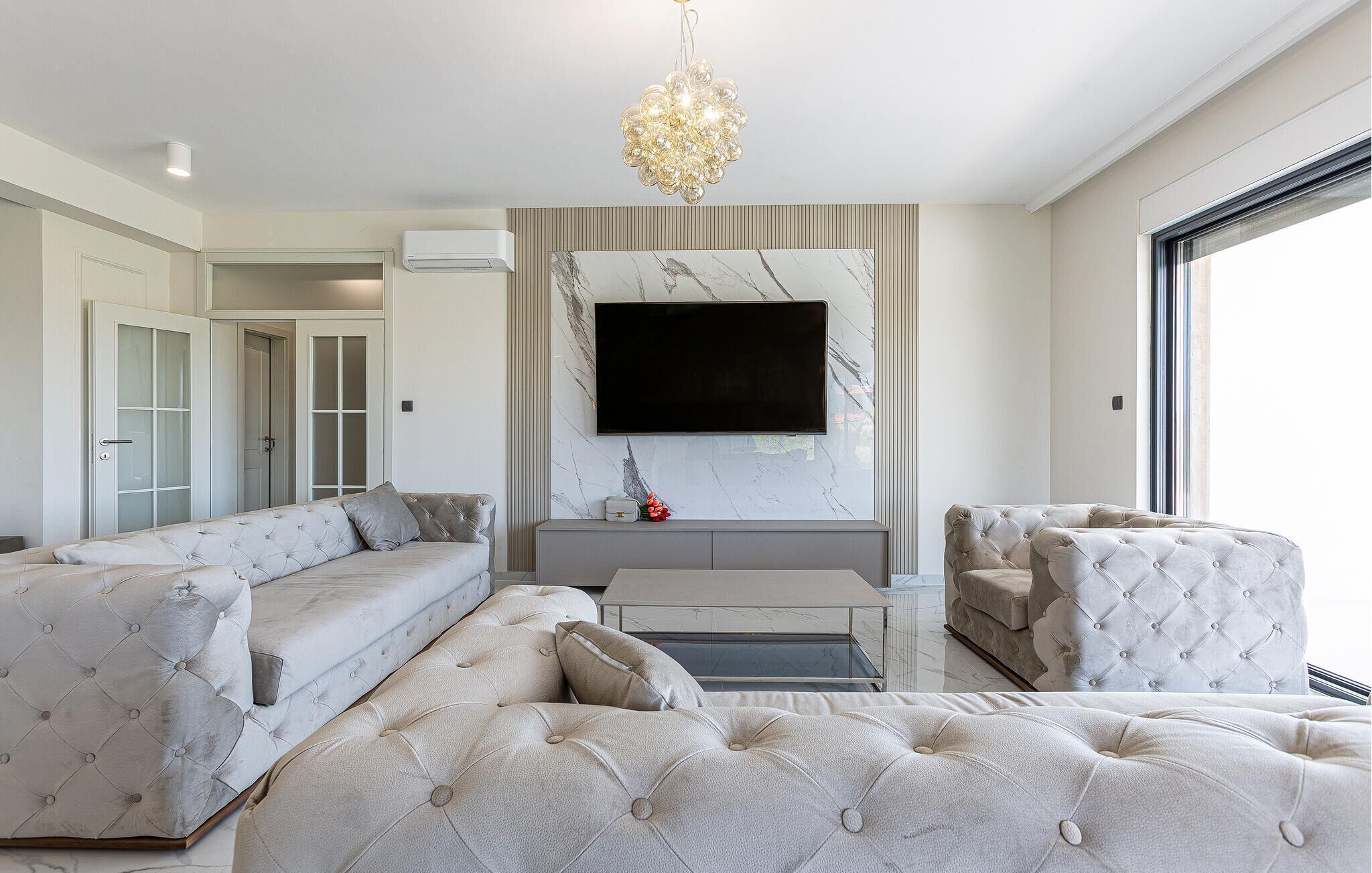 A modern living room with a large sectional sofa, a tufted ottoman, a flat-screen TV mounted on a marble wall, and a decorative chandelier.