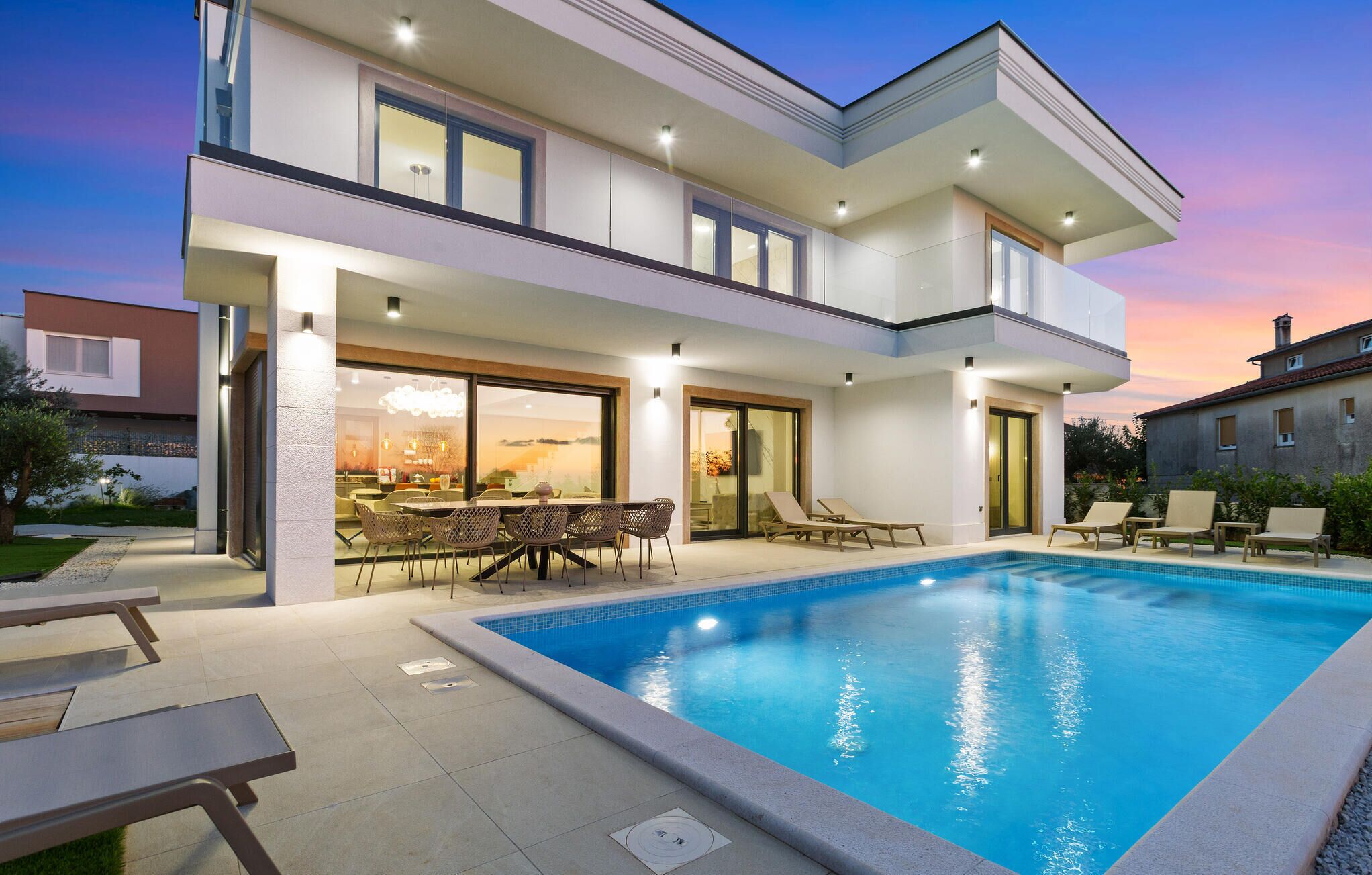 A modern two-story house with a swimming pool, outdoor seating, and a large glass door leading to an interior dining area.