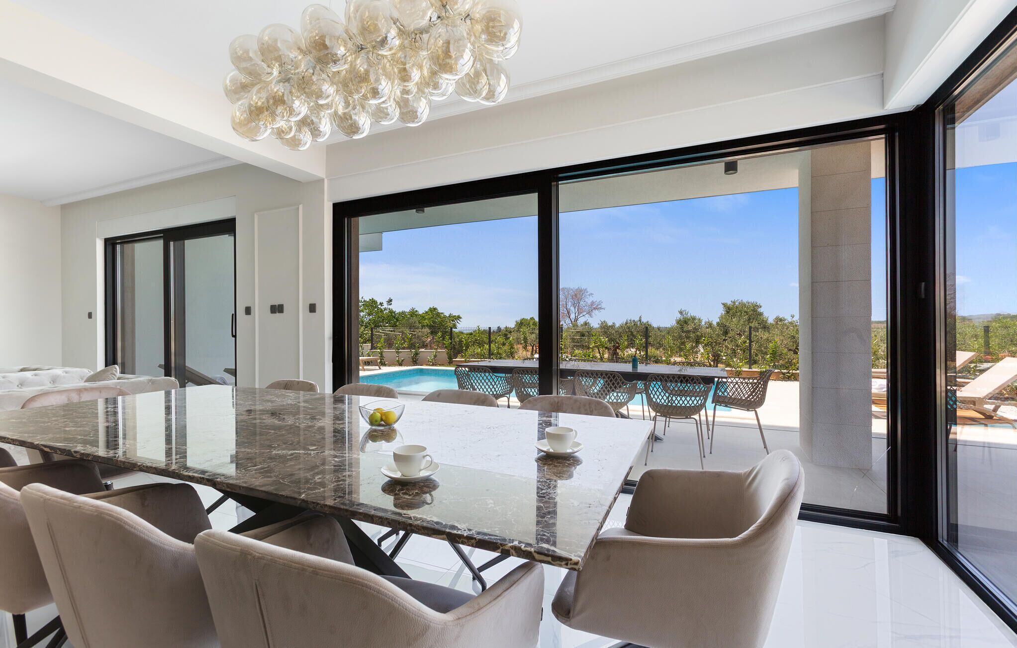 A modern dining area with a glass table and chairs, large windows, and a view of a pool and greenery.