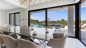 A modern dining area with a glass table and chairs, large windows, and a view of a pool and greenery. - Stunning home in Galilzana (Galilzana)