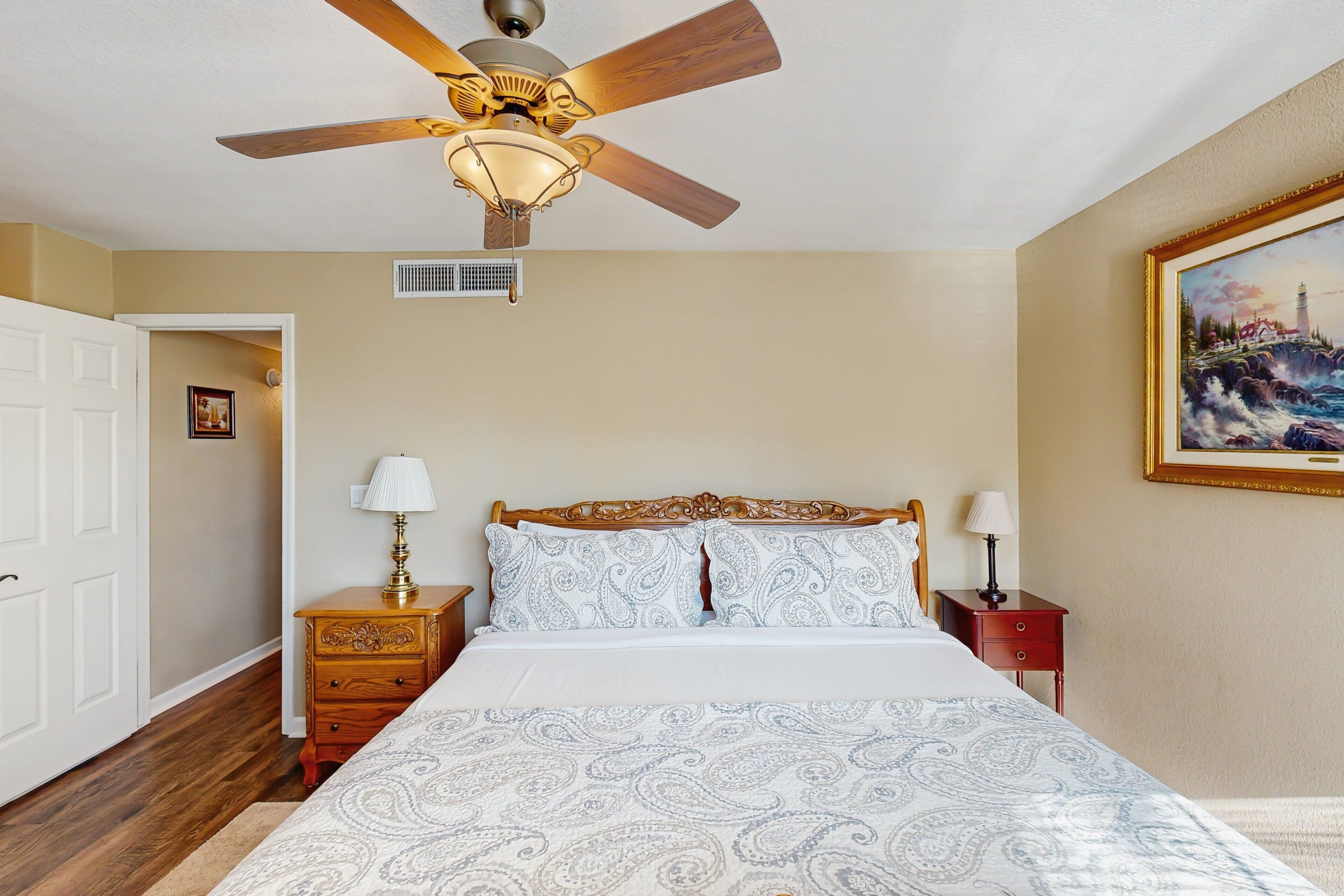 Quarto com cama, cabeceira de madeira, duas mesas de cabeceira com abajures, ventilador de teto, um quadro na parede e uma porta que dá para outro cômodo.