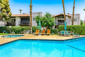 A swimming pool with lounge chairs and umbrellas, surrounded by palm trees and a building with balconies. - Modern condo with mountain view, private balcony, pools (Palm Springs)