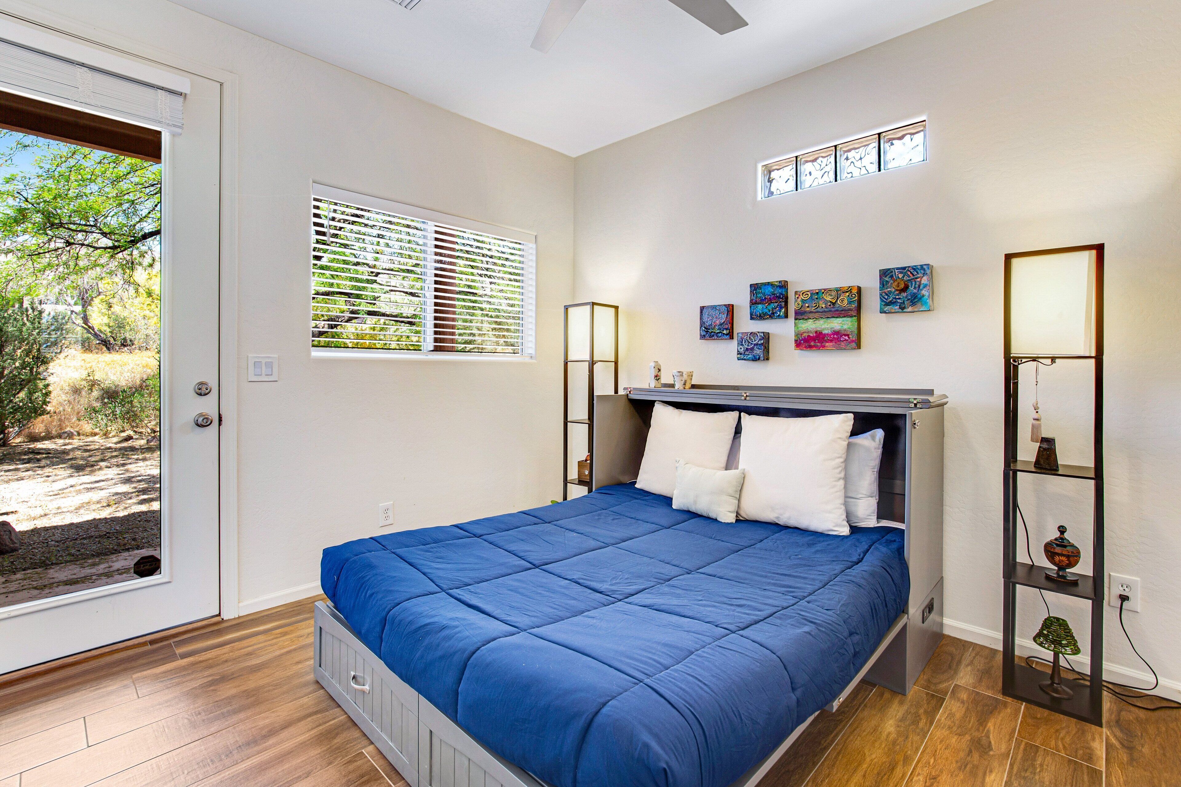 Une chambre à coucher avec un lit, une fenêtre avec des stores, une étagère fixée au mur et des œuvres d’art.