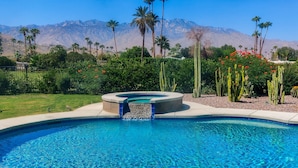 A swimming pool with a waterfall feature, surrounded by palm trees and cacti, with mountains in the background. - Mid-century modern retreat with pool/spa & firepit - golf & mtn views (Palm Springs)