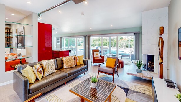 A modern living room with a leather sofa, glass shelving, a fireplace, and a view of the outdoors. - Mid-century modern retreat with pool/spa & firepit - golf & mtn views (Palm Springs)