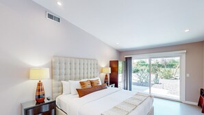 A bedroom with a large bed, two bedside tables, a window with a view of a garden, and a ceiling vent. - Mid-century modern retreat with pool/spa & firepit - golf & mtn views (Palm Springs)