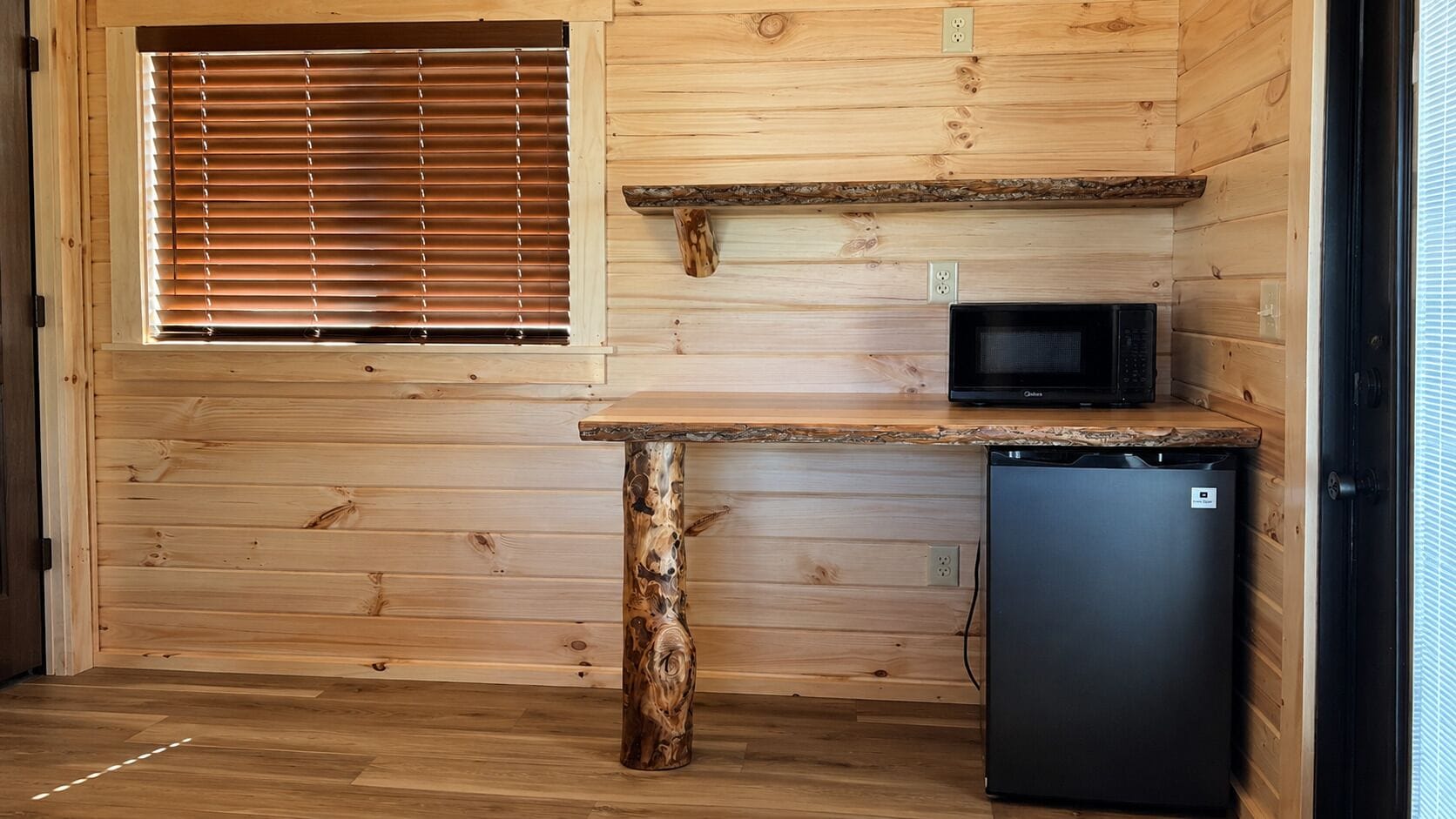 Eine kompakte Küche mit Holzwänden, einer integrierten Mikrowelle, einem kleinen Kühlschrank und einem Fenster mit Jalousien.