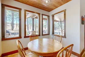 A dining area with a round table and four chairs, large windows with a view of a forest, and a wooden ceiling.