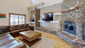 A living room with a stone fireplace, leather sofa, and a flat-screen TV mounted on a stone wall. - Exquisite home with mountain views, fireplaces, game room & private Hot tub (Flagstaff)