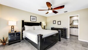 Une chambre à coucher comprenant un lit, une table de chevet, une commode, un ventilateur de plafond et une salle de bain visible à travers une porte ouverte.