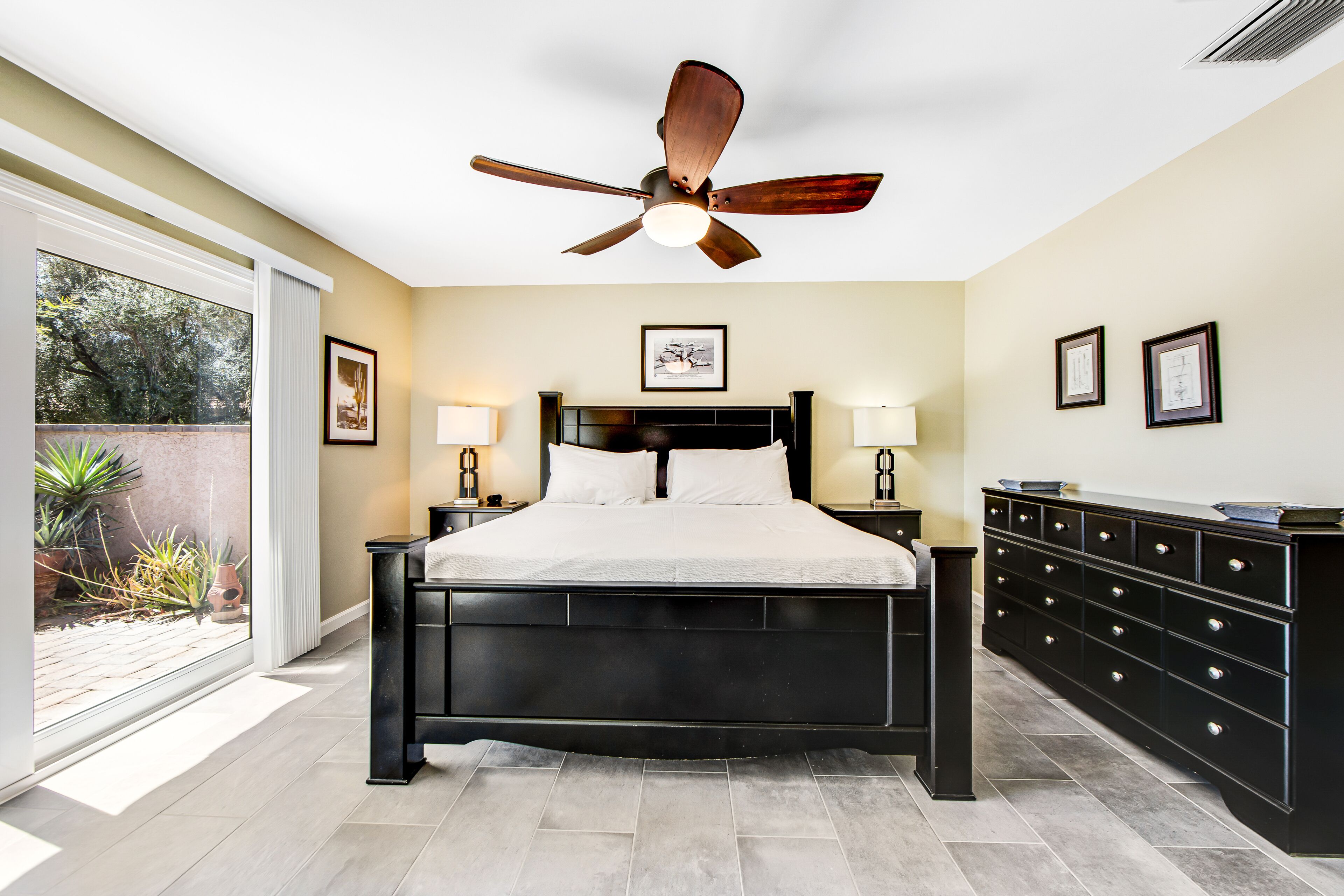 A bedroom with a bed, nightstands, a dresser, a ceiling fan, and a large window with a view of greenery.