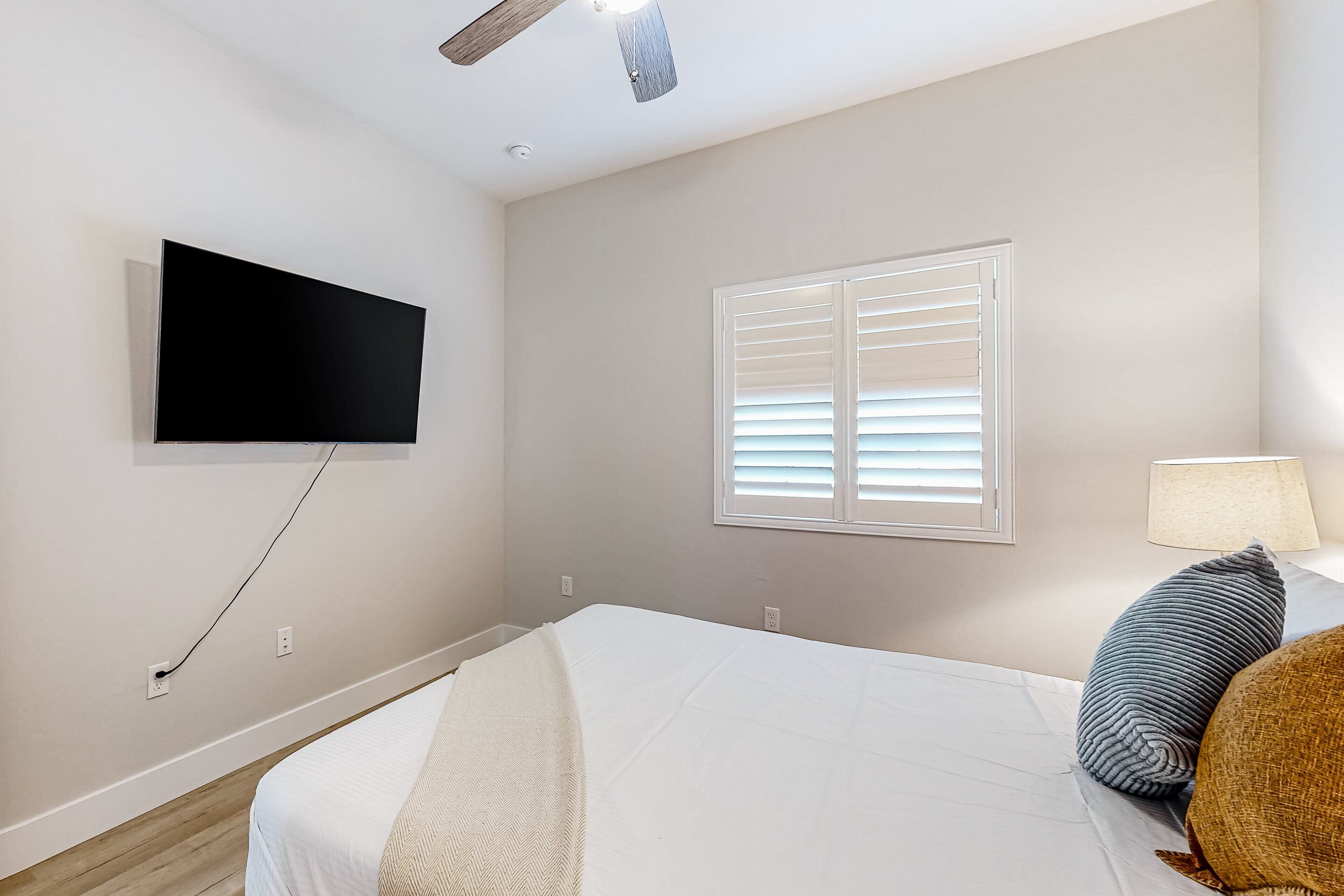 Une chambre à coucher comprenant un lit, un téléviseur à écran plat fixé au mur, une fenêtre avec des stores et un ventilateur de plafond.