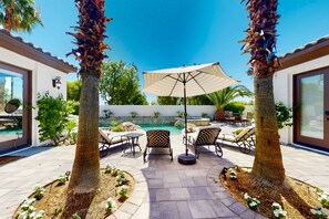 A patio area with a pool, palm trees, outdoor furniture, and a covered umbrella.