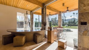 Une cuisine moderne dotée d’un grand îlot central, d’un plafond en bois et de grandes fenêtres offrant une vue sur les palmiers et les montagnes.