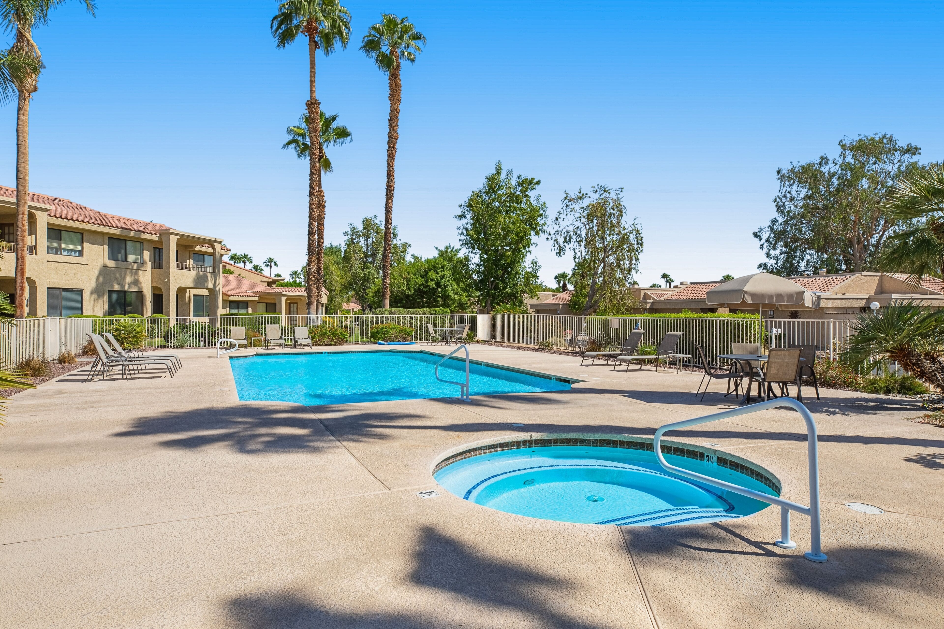 A swimming pool with lounge chairs and a small hot tub, surrounded by palm trees and a fence.