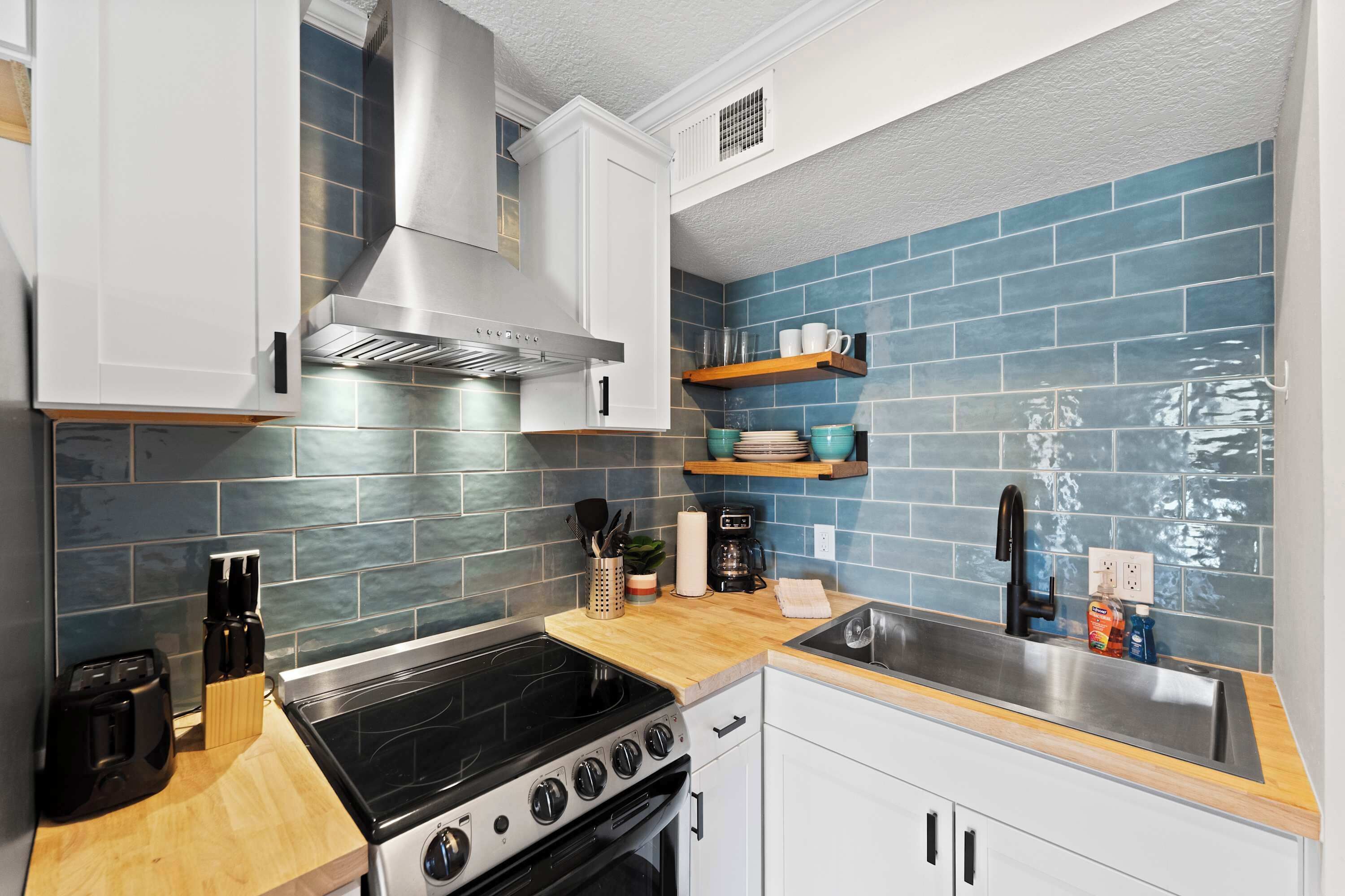 A kitchen with white cabinets, a stainless steel range hood, a black stove, and a blue-tiled backsplash.