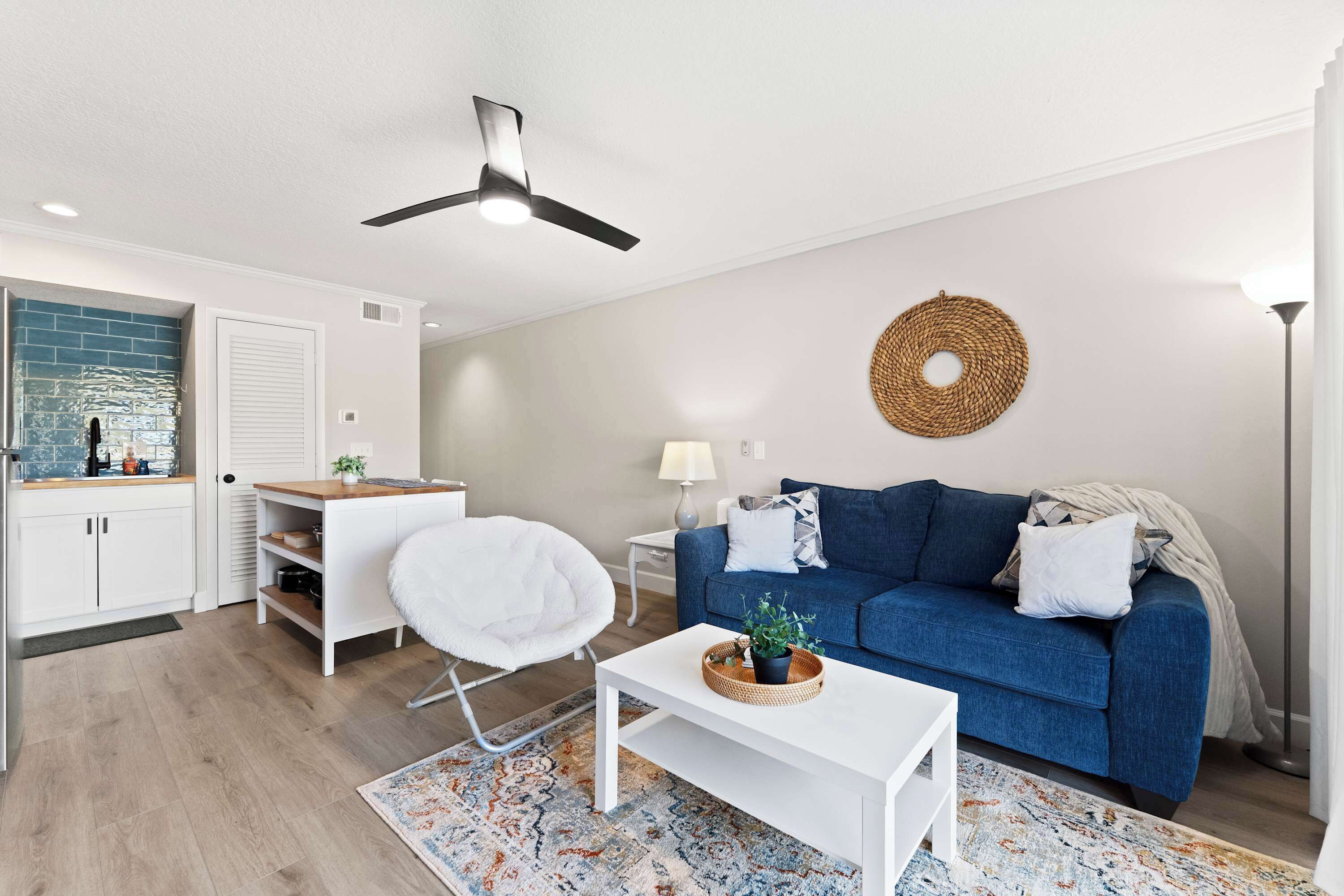 En moderne stue med en blå sofa, en hvid lænestol og et hvidt sofabord. Der er en ventilator i loftet, et vævet gobeleng og en lampe på et sengebord.