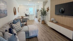 A modern living room with a sectional sofa, a flat-screen TV mounted on a wooden wall, a kitchen in the background, and a staircase leading to another level. - Modern Coastal Townhouse Near Beach & DT - St. Augustine (St. Augustine)