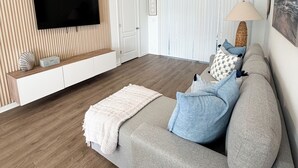 A modern living room with a flat-screen TV mounted on a wooden wall, a grey sofa with blue cushions, a woven floor lamp, and a large window with blinds. - Modern Coastal Townhouse Near Beach & DT - St. Augustine (St. Augustine)