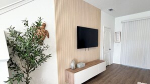 A modern living room with a flat-screen TV mounted on a wooden panel wall, a wooden bench with decorative items, a potted olive tree, and a cozy area rug. - Modern Coastal Townhouse Near Beach & DT - St. Augustine (St. Augustine)