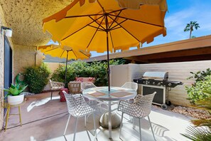 Una terrazza esterna con un tavolo rotondo, sedie bianche, ombrelloni gialli e un barbecue.