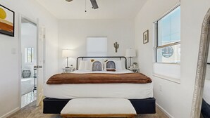 Et soveværelse med seng, sengeborde, lampe, et vindue, en loftsvifte og en dør.