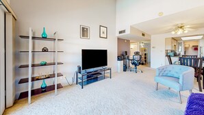 A living room with a flat-screen TV, a glass coffee table, a blue armchair, and a bookshelf with decorative items. - Gorgeous condo with hot tub, pool, tennis court, & grilling area - lake access (Phoenix)