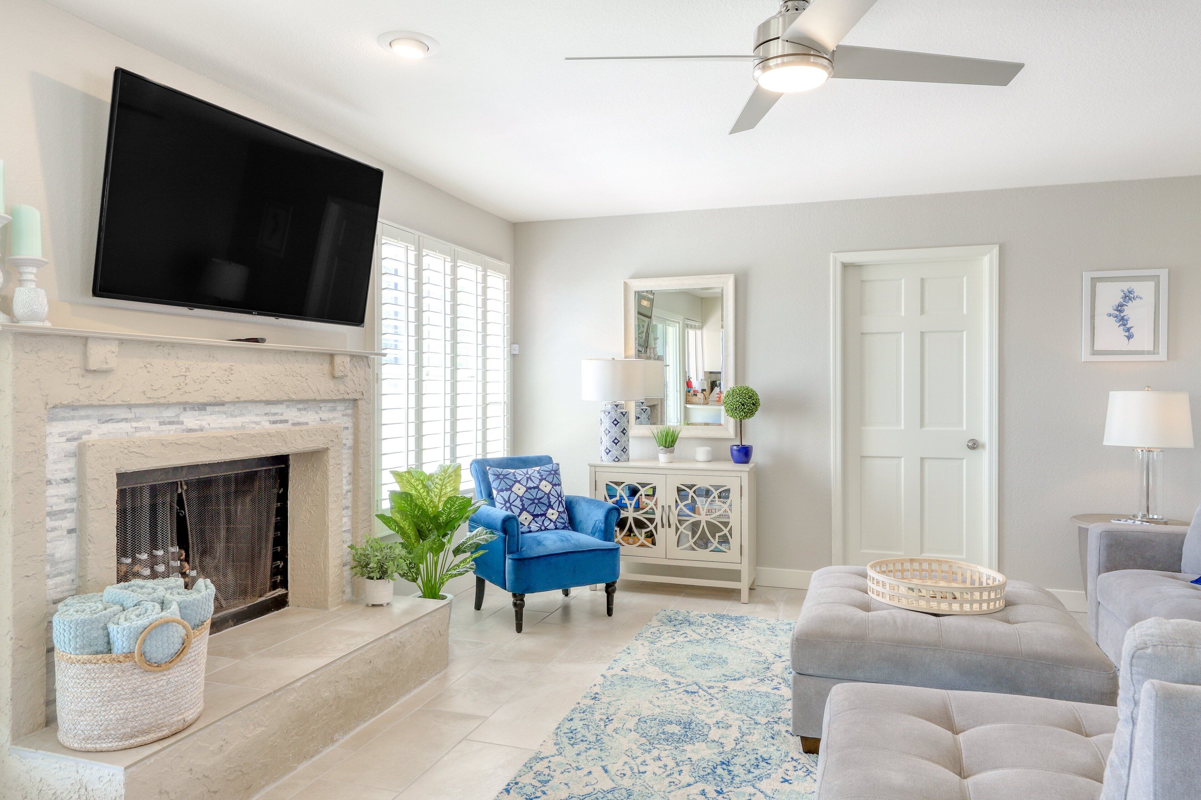A living room with a flat-screen TV mounted above a fireplace, a ceiling fan, a blue armchair, a grey sofa, and a patterned rug.