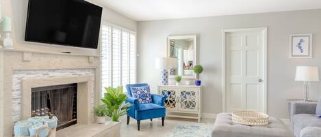 A living room with a flat-screen TV mounted above a fireplace, a ceiling fan, a blue armchair, a grey sofa, and a patterned rug.