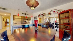 Ruime eetruimte met een grote houten tafel, een moderne keuken op de achtergrond en een rustieke houten kast met planken.