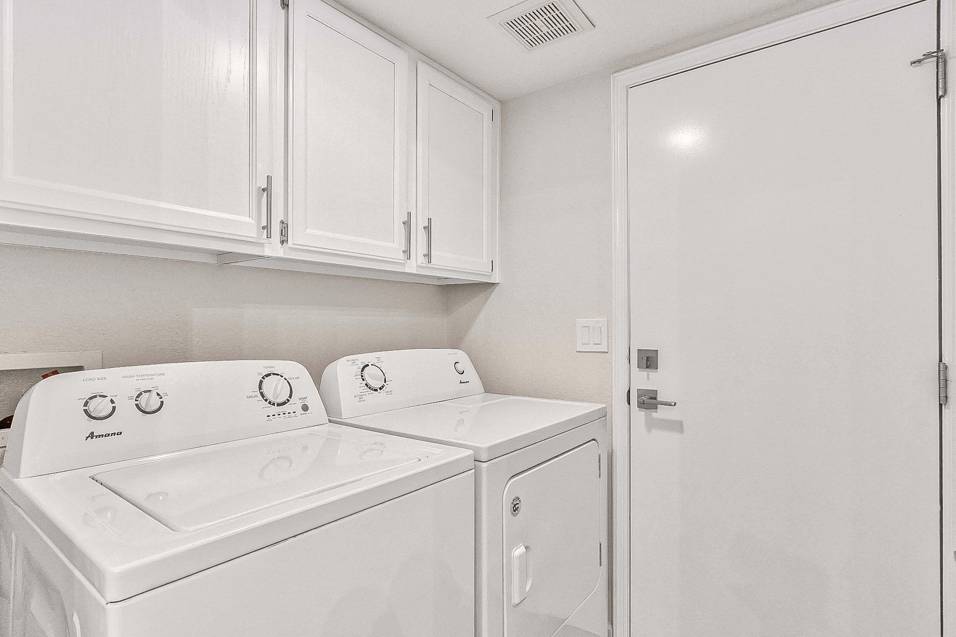 A laundry room with two white washing machines, a door, and a vent on the ceiling.