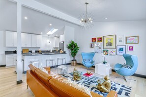 Ein modernes Wohnzimmer mit einer braunen Ledersofa, einem Glastisch und einem blauen Sessel. Im Hintergrund ist eine Küche mit weißen Schränken und einer Kochinsel zu sehen.
