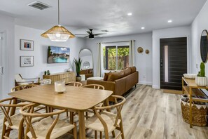 Amplia sala de estar con una mesa de comedor de madera, asientos cómodos, un televisor grande y vistas al exterior.