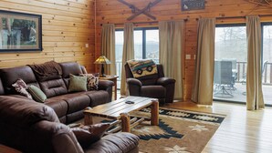 Ein gemütliches Blockhaus-Interieur mit Ledersofa, Ohrensessel und Esstisch. Ein gerahmtes Bild hängt an der Wand, und ein Schild zeigt den Namen der Lodge.