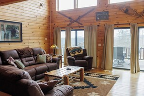 Ein gemütliches Blockhaus-Interieur mit Ledersofa, Ohrensessel und Esstisch. Ein gerahmtes Bild hängt an der Wand, und ein Schild zeigt den Namen der Lodge.