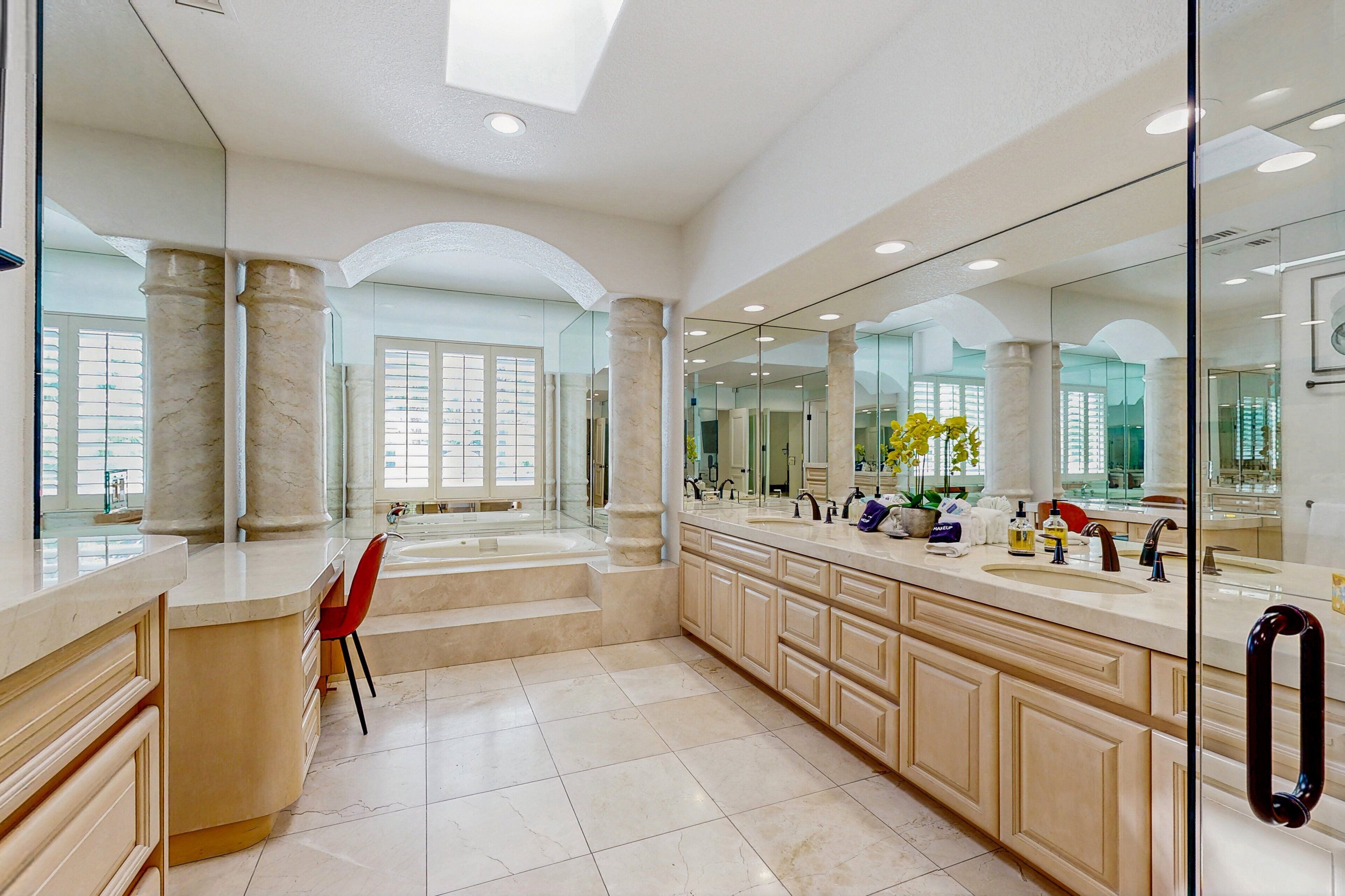 A spacious bathroom with a large mirror, two sinks, a red chair, and a potted plant.