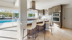 Eine moderne Küche mit einer großen Kochinsel, Barhocker, einem eingebauten Backofen und Blick auf einen Swimmingpool.