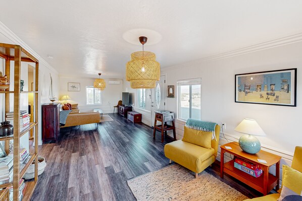 A spacious living room with a wooden floor, a yellow sofa, a bookshelf, and a large window. - Cacti Sanctuary with private hot tub & firepit near Joshua Tree (Twentynine Palms)