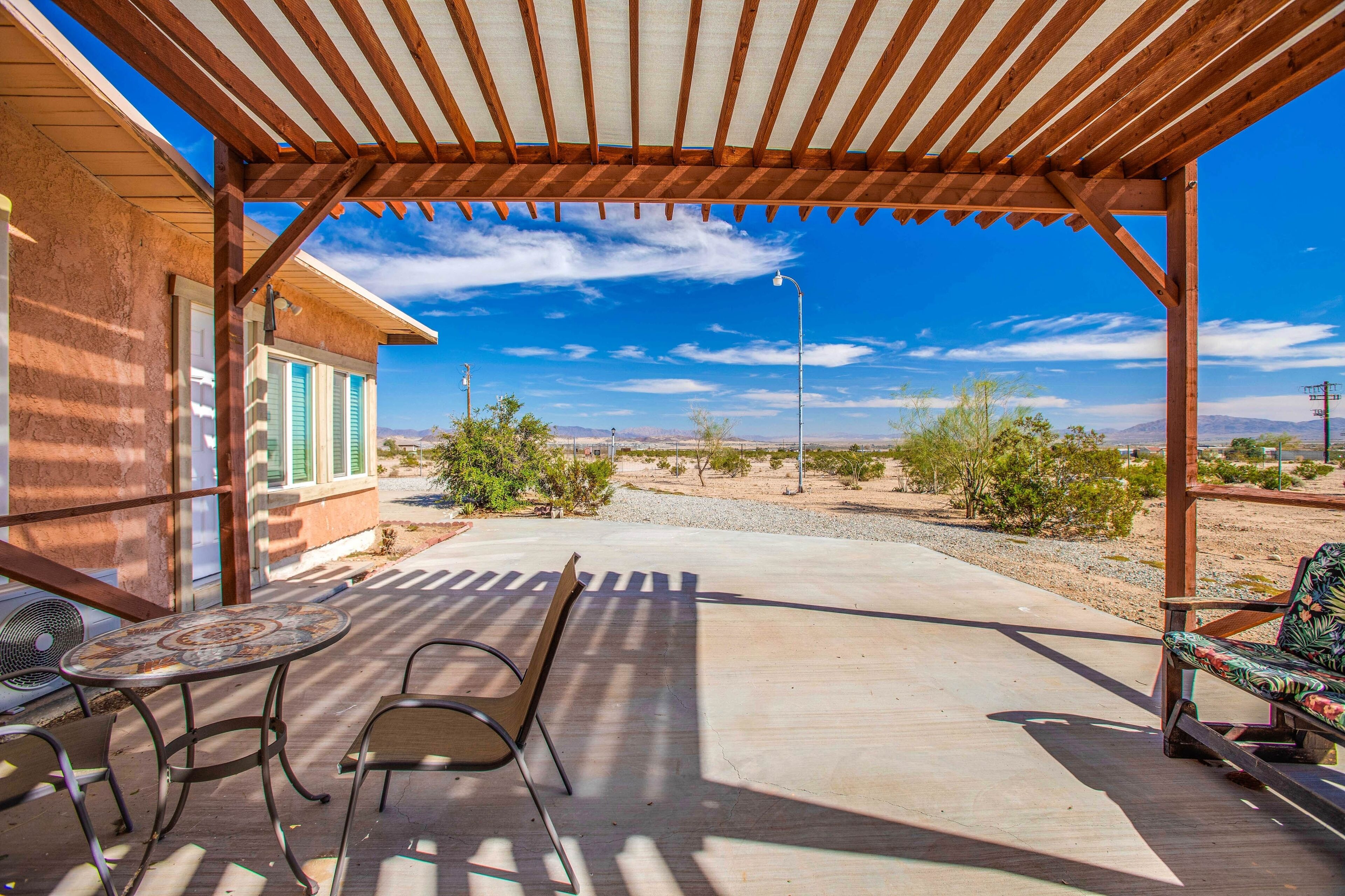 A covered patio with a table and chairs, a view of a desert landscape, and a building with green shutters.