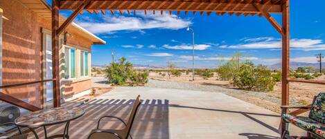 A covered patio with a table and chairs, a view of a desert landscape, and a building with green shutters.