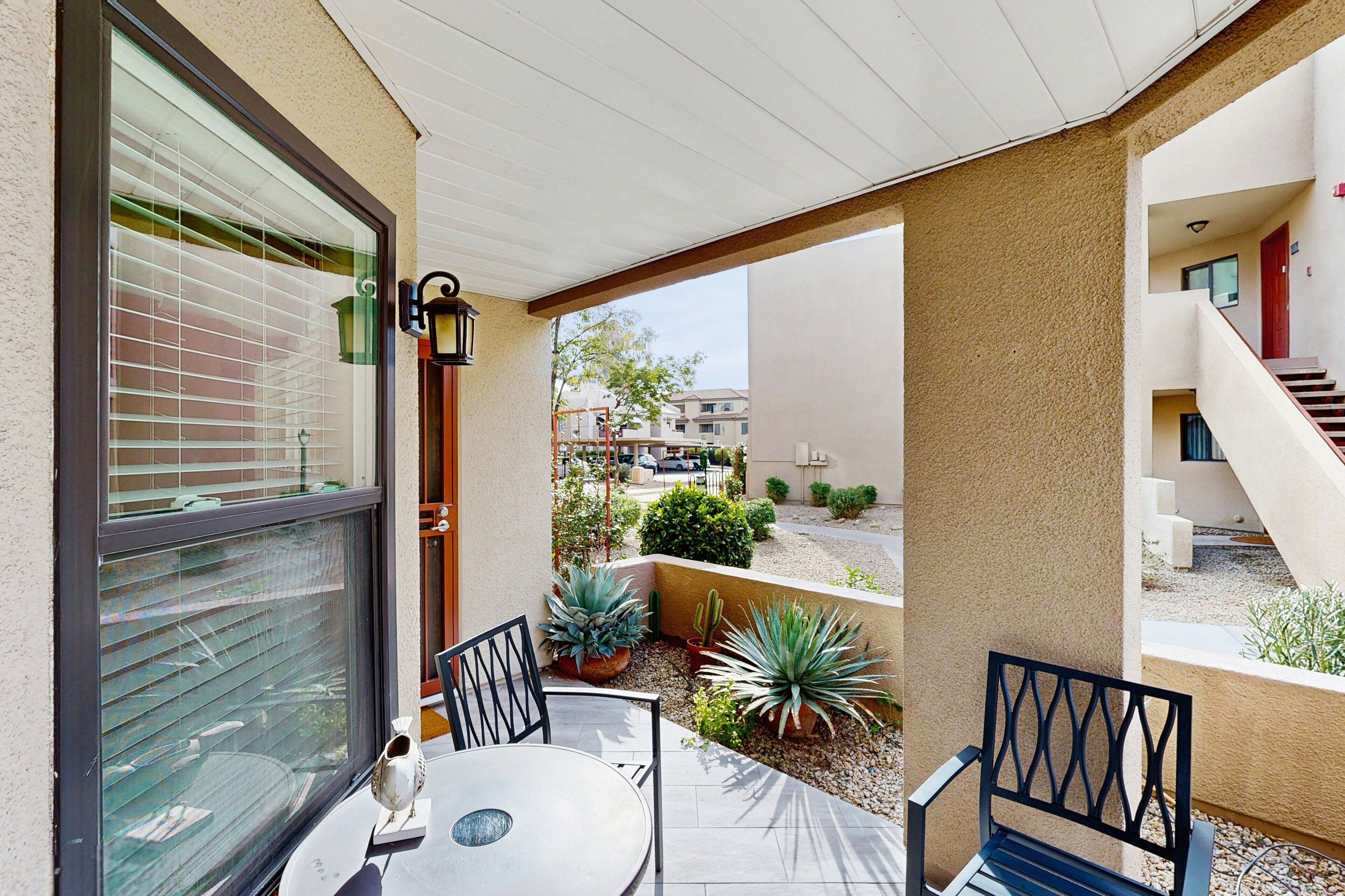 A covered patio with a table and chairs, potted plants, and a view of the street and buildings.