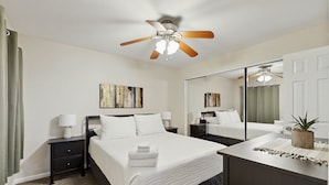 Une chambre d’hôtel avec un lit, une table de chevet, une commode, un ventilateur de plafond, un miroir et un tableau au mur.