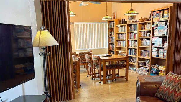 Una sala de estar con televisor, una mesa de centro con un libro, un sofá, un área de comedor con sillas y una estantería llena de libros y juegos.