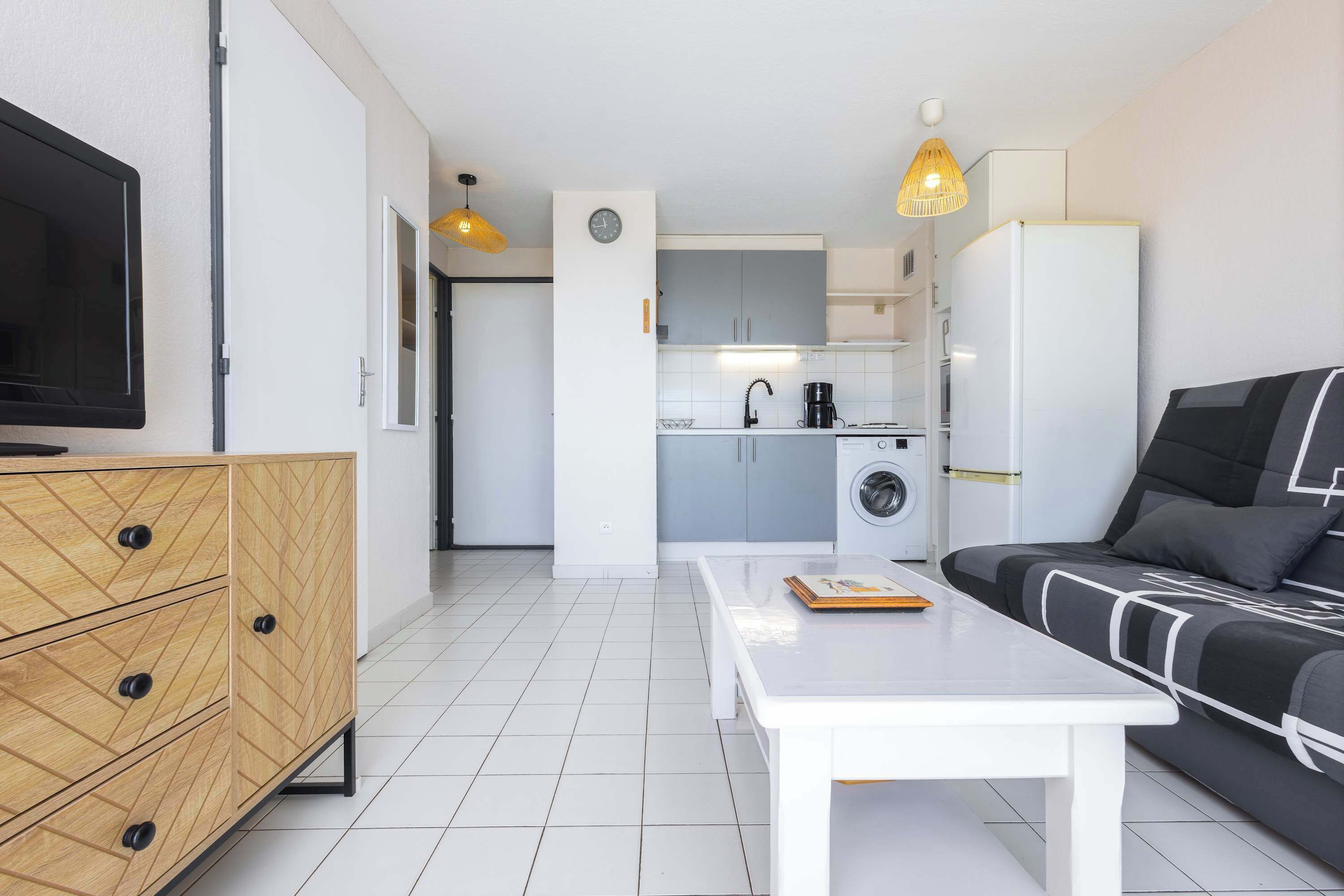 Un appartement moderne doté d’une télévision, d’une commode en bois, d’une table blanche, d’un canapé, d’une cuisine avec un lave-linge et d’une horloge murale.