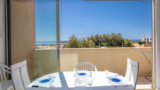 Un balcó amb taula preparada per dinar, amb vista a una platja i arbres.
