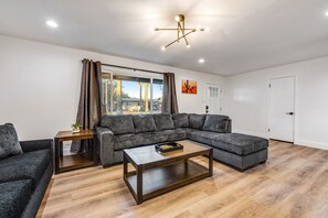 A modern living room with a sectional sofa, a coffee table, and a large window with curtains. - Stylish desert oasis with fast WiFi, fenced yard, & screened patio (Scottsdale)