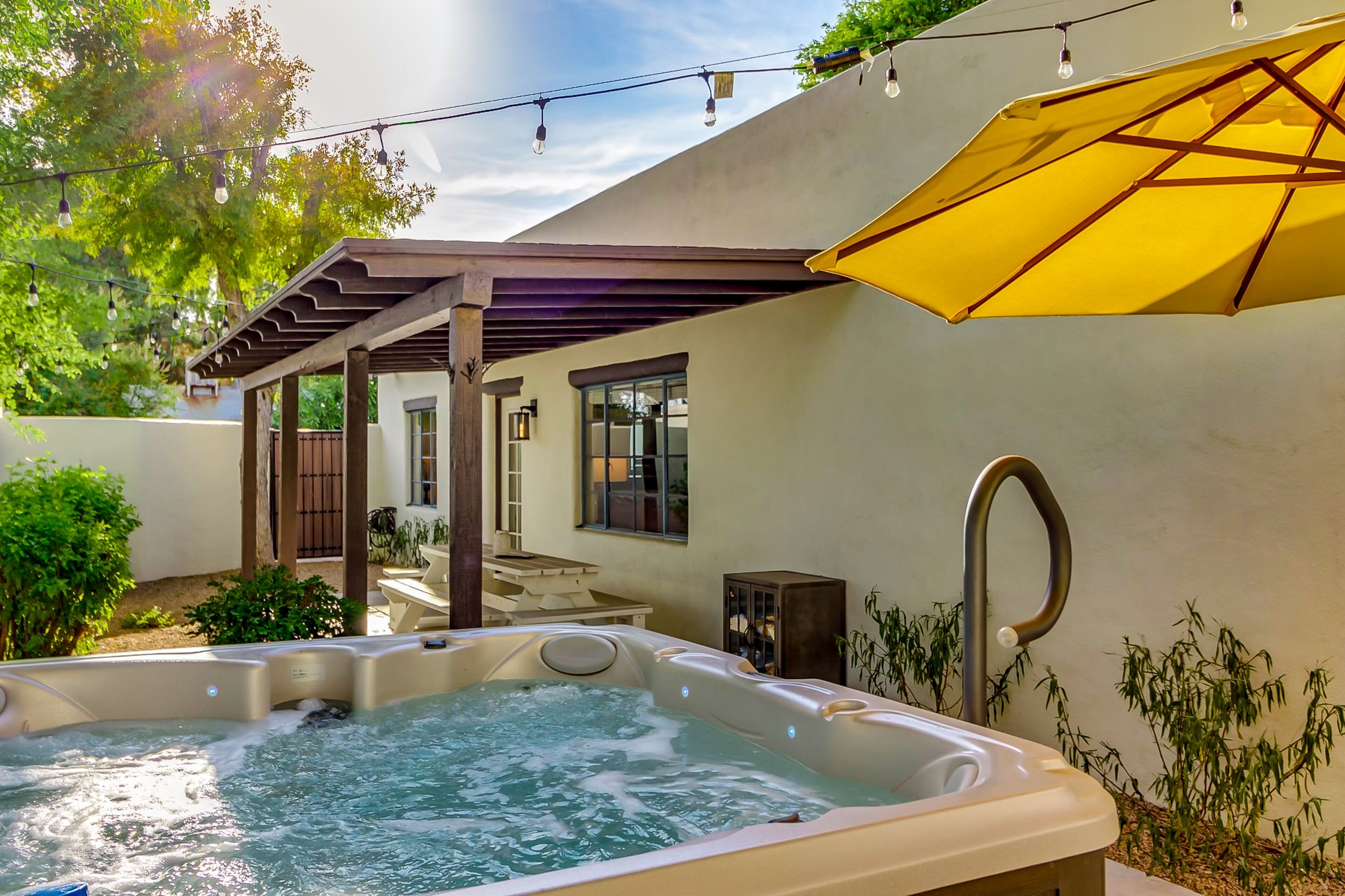 A hot tub with a pergola and a yellow umbrella, surrounded by greenery and a building with windows.