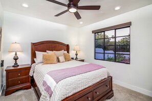 A bedroom with a wooden bed, two bedside tables with lamps, a ceiling fan, and a window with a view of greenery. - Escape to This Charming Single-Level Home in Scottsdale, AZ (Scottsdale)